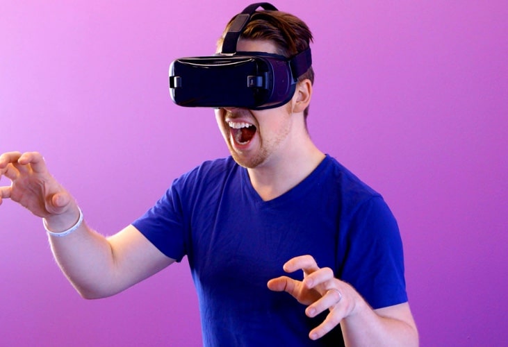 a man who has a short brown hair and wears blue t-shirt and wears virtual reality glassess and plays a video game